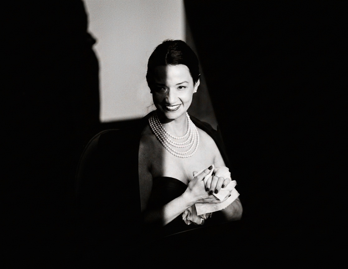 A woman in pearls smiling softly in dramatic chiaroscuro light